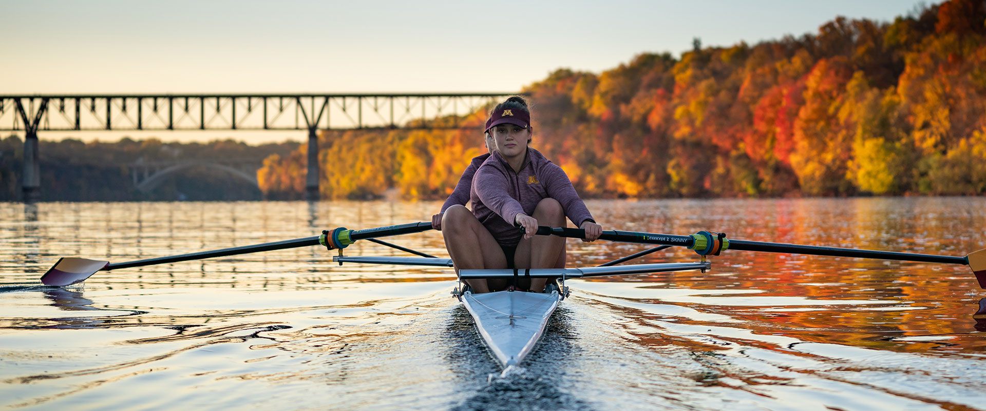 Women rowing on river with bridge and trees in background